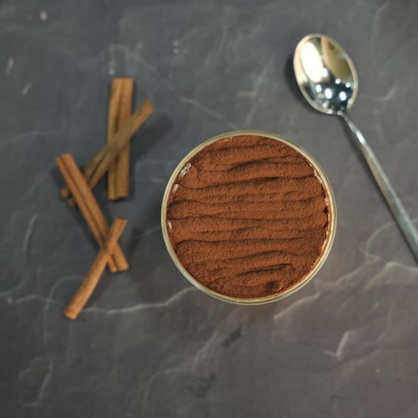 Overhead view of Grano's tiramisu in a glass, dusted with cocoa, alongside cinnamon sticks and a spoon.